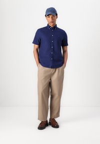 Camicia blu navy a maniche corte con bottoni, pantaloni beige a gamba larga, scarpe marroni scure e un berretto azzurro chiaro, con un logo sottile sulla camicia.