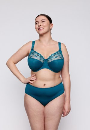 Vrouw poseert zelfverzekerd in een teal kanten en stoffen beha met bijpassend hoog opgesneden ondergoed tegen een effen lichte achtergrond.
