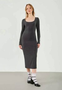 Robe ajustée à manches longues, couleur gris foncé, en tissu doux, avec un décolleté carré et une longueur au genou. Portée avec des talons noirs et des chaussettes rayées.