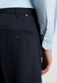 Granatowe spodnie z tylną kieszenią, zapięciem na guzik i małą pomarańczową metką. Widoczny mankiet jasno-niebieskiej koszuli obok spodni.