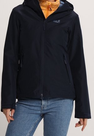 Sort outdoorjakke med lynlås og hætte over orange fleece, båret af en person i blå jeans, vist fra nakke til hofte.