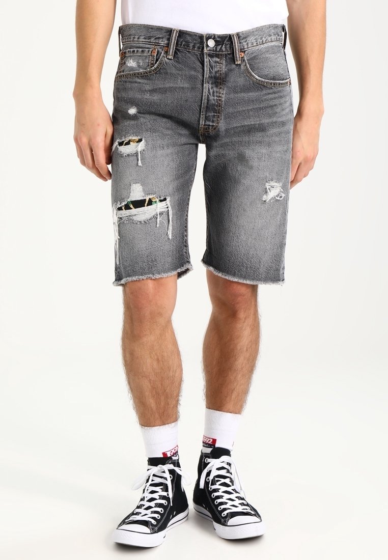 Hombre vistiendo pantalones cortos de mezclilla negros desgastados y rasgados, calcetines blancos y zapatillas altas negras frente a un fondo liso.