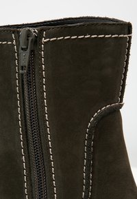 Detalle de una bota de ante oscuro con un zipper negro visible y costuras de contraste blancas a lo largo de los bordes y alrededor del zipper.