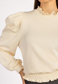 Crème-kleurige sweatshirt met een volants-halslijn en poofy mouwen. Bevat smockdetails in de tailleband en heeft een gladde, zachte textuur.