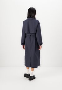 Cappotto lungo navy con vita aderente, maniche larghe e schiena spaccata. Abbinato a scarpe nere robuste e calzini bianchi. Tessuto dalla texture liscia.