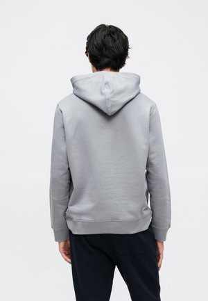 Άνδρας που φοράει ανοιχτό γκρι φούτερ με κουκούλα και σκούρο παντελόνι, όρθιος με την πλάτη προς την κάμερα σε λευκό φόντο.