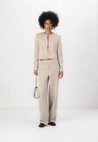 Giacca cropped beige decorata con perline, maniche lunghe, scollatura rotonda e zip, abbinata a pantaloni beige a gamba larga e una piccola borsa.