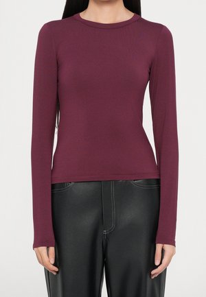 Frau trägt ein eng anliegendes langärmeliges dunkelviolettes Oberteil und hochtaillierte schwarze Lederhosen mit sichtbarer weißer Naht.