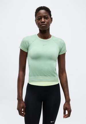 Vrouw draagt een mintgroen sporttopje met korte mouwen en zwarte Nike legging, staand tegen een effen lichte achtergrond.