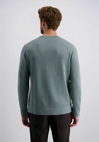Herren Pullover mit langen Ärmeln aus einem weichen, strukturierten, gedämpften grünen Stoff mit Rundhalsausschnitt und geradem Saum, getragen über dunklen Hosen.