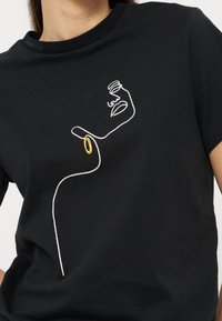 Μαύρο μπλουζάκι με ένα λευκό σχέδιο μιας συνεχούς γραμμής που απεικονίζει το πρόσωπο και τον λαιμό μιας γυναίκας, με ένα χρυσό κρίκο ως σκουλαρίκι.