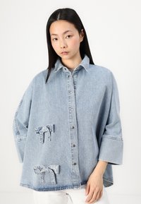 Ljusblå denim skjorta i oversize med spetsig krage, tryckknappar och två dekorativa rosettaccent på framsidan. Långa ärmar med manschetter.