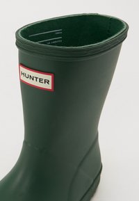 Bottes de pluie en caoutchouc vertes avec un dessus arrondi, un intérieur texturé et une étiquette avant avec "HUNTER" en rouge et blanc. La semelle est striée.