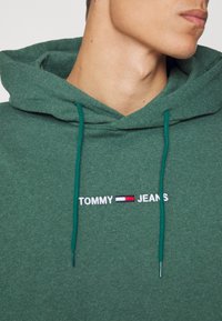 Grön hoodie med mjuk textur, med dragsko i matchande färg och "TOMMY JEANS" broderat i vitt och rött.