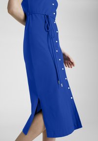 Blå klänning med knappar längs framsidan, knuten i midjan med ett matchande bälte. Har en sidofrans och en slät tygtextur.