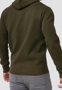 Olivgrüner Hoodie mit strukturiertem Strick, ausgestattet mit Kapuze und gerippten Bündchen und Saum, kombiniert mit hellgrauen Hosen.