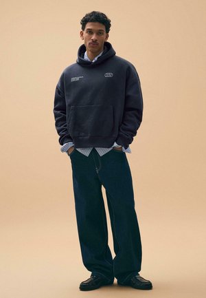 Man met krullend haar draagt een zwarte hoodie over een gestreept shirt, losse donkere jeans en zwarte schoenen, staand met handen in de zakken tegen een beige achtergrond.