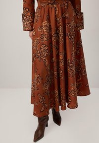 Falda floral marrón con patrones florales negros llamativos, diseño fluido, tela ligera, cintura ceñida, combinada con botas de tacón puntiagudas marrones.