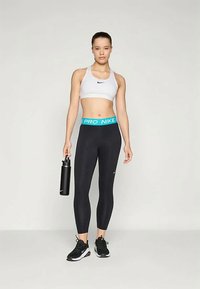 White sports bra with Nike logo, black leggings with aqua "PRO NIKE" waistband, holding a black water bottle, wearing black sneakers.