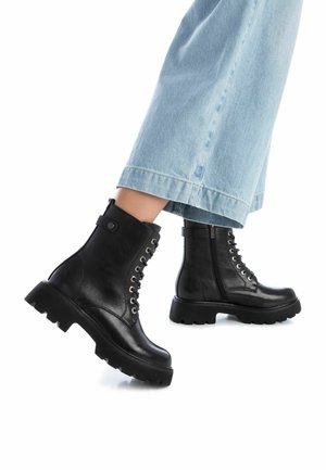 Stivali alla caviglia in pelle nera con lacci frontali, occhietti metallici, zip laterale e suola in gomma spessa, abbinati a jeans ampi blu chiaro.