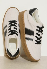 Balti odiniai sportiniai bateliai su juodais akcentais ir raišteliais, tekstūruota gumine padu ir dviem juodomis juostomis šonuose.