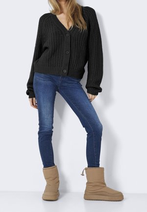 Kvinde iført en sort chunky strikket cardigan, blå skinny jeans og tan isolerede vinterstøvler, der står mod en hvid baggrund.