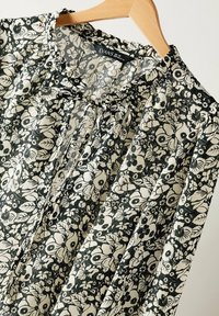 Blomstret bluse med sort baggrund, prydet med hvide blomstermønstre, plisseret design og rund halsudskæring. Mærket viser størrelse UK 10.