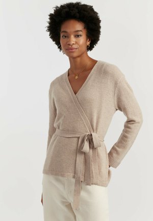 Pull beige cache-cœur en tissu doux, doté d'un décolleté en V profond, de manches longues et d'une ceinture nouée à la taille. Design texturé et confortable.