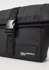 Borsa pieghevole nera realizzata in materiale liscio, con una fibbia in plastica e una tracolla con logo, che presenta un'etichetta con logo bianco in evidenza.
