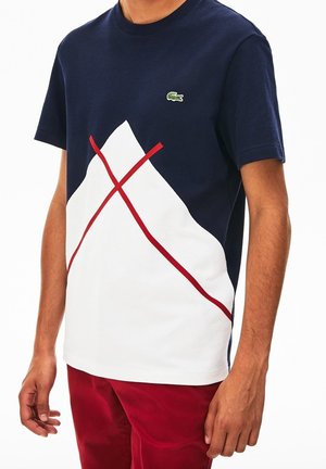 Mand iført mørkeblå og hvid t-shirt med røde krydslinjer og Lacoste-logo, kombineret med røde bukser, stående med afslappede arme.