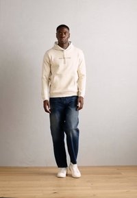 Felpa beige con tasca frontale e coulisse, abbinata a jeans blu e sneakers bianche; posizionata su uno sfondo chiaro e uniforme.
