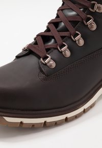 Bota de senderismo de cuero marrón con acabado texturizado, amplios cordones marrones, ojetes de metal y suela de goma color crema con un patrón de relieve.