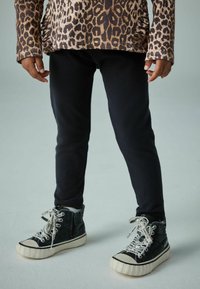 Schwarze, enganliegende Sweatpants kombiniert mit hochgeschnittenen schwarzen Schuhen, die auffällige weiße Schnürsenkel haben. Das Oberteil hat ein Leopardenmuster.