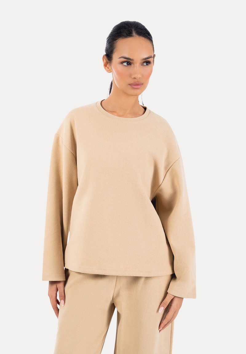 Donna con capelli scuri, indossa una maglia beige a maniche lunghe e pantaloni abbinati, in piedi con le mani rilassate, guarda verso destra su uno sfondo bianco.