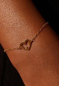 Gouden armband met een hartvormige hanger waarin een vlinder is uitgesneden, bevestigd aan een delicaat ketting, rustend op een bruine huid.