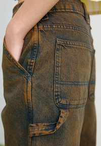 Pantaloni in denim con un lavaggio marrone e blu sbiadito, dotati di tasche laterali e dettagli di cucitura visibili, che mostrano un tessuto testurizzato e una vestibilità rilassata.