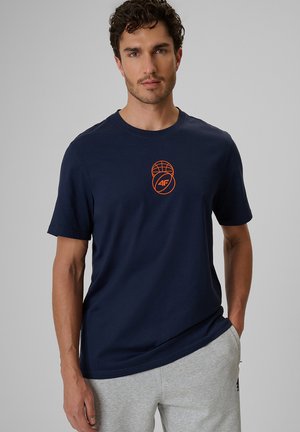 Mężczyzna z krótkimi kręconymi włosami, ubrany w granatowy t-shirt z pomarańczowym nadrukiem i jasnoszare spodnie dresowe, stoi na jednolitym tle.