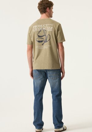 Olijfgroene katoenen T-shirt met grafische tekst en bloemenontwerp op de achterkant, gecombineerd met blauwe spijkerbroek en bruine schoenen.