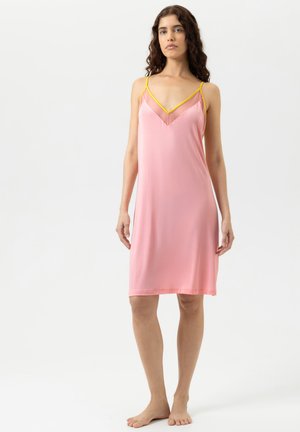 Giovane donna con capelli ricci che indossa un vestito rosa con bordo giallo, in piedi a piedi nudi contro uno sfondo bianco semplice.