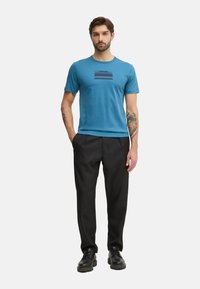 Camiseta corta de manga azul con un diseño de logotipo a rayas horizontales; combinada con pantalones negros y zapatos negros de cordones, con un ajuste relajado.