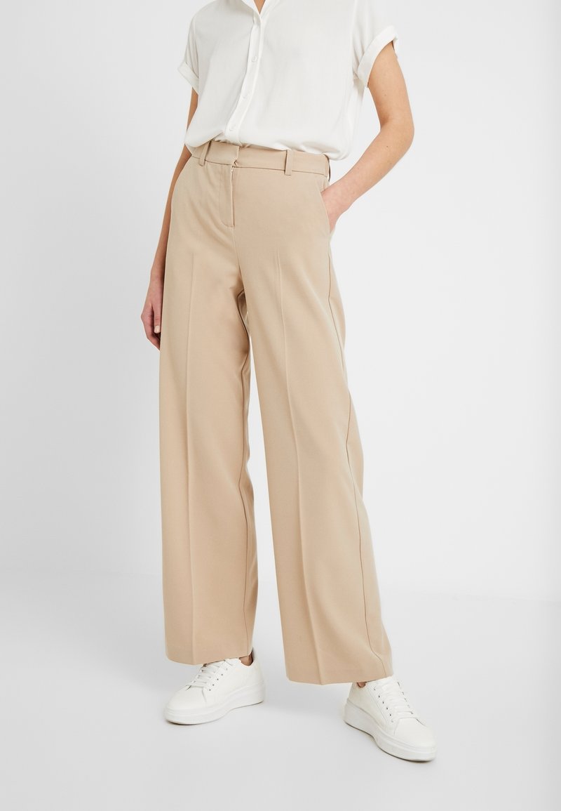 Personne portant un pantalon large beige, une chemise blanche à manches courtes boutonnée et des baskets blanches, avec une main dans la poche, debout sur un sol blanc.