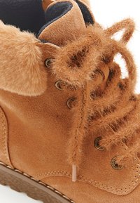 Tan suède laars met een zachte faux-fur kraag, textuurveters, metalen oogjes en een bruine rubberen zool. Close-up kijk benadrukt materiaaldetails.