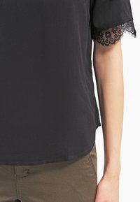 Svart kortärmad topp i mjukt material, med spetsdetaljer längs ärmarna, avslappnad passform och rundad nederkant. Kombineras med olivgröna byxor.