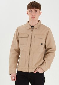 Beige zip-up jakke laget av slitesterk stoff, med en spiss krage, to frontlommer og en liten logo-detalj på brystet.