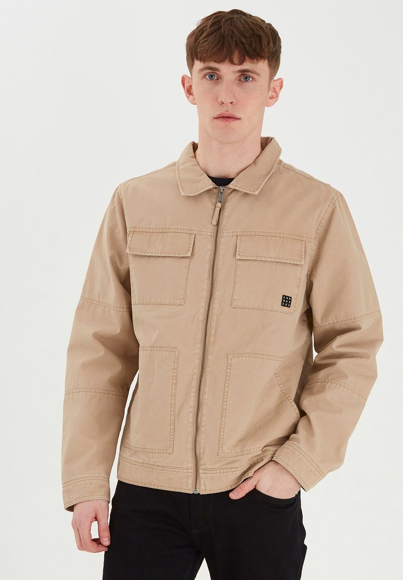 Beige zip-up jakke laget av slitesterk stoff, med en spiss krage, to frontlommer og en liten logo-detalj på brystet.