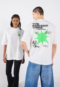 White oversized t-shirt with green star and graphic print on front and back. Styled with black pants and blue wide-leg jeans.