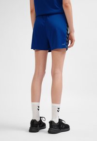 Blauwe sportshorts met een comfortabele elastische tailleband, voorzien van een chevron-patroon langs de zijkanten. Gecombineerd met zwarte sneakers en witte sokken.
