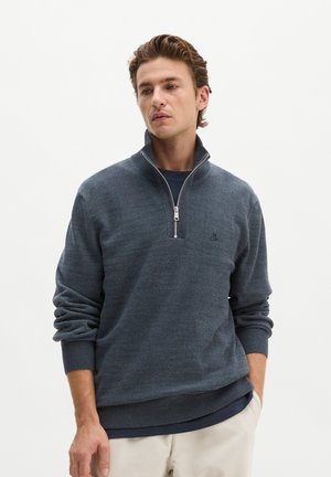 Man with wavy brown hair wearing a dark blue zip-up sweater and cream pants, standing with one hand in pocket, looking down and to the side.