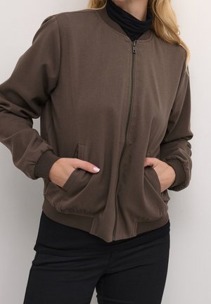 Frau trägt eine braune Bomberjacke mit Reißverschluss über einem schwarzen Rollkragenpullover, die Hände in den Jackentaschen, vor einem schlichten hellen Hintergrund.