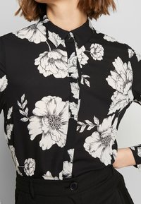Blouse noire boutonnée avec un grand imprimé floral blanc, portée rentrée dans un pantalon noir, montrant le col et le milieu du corps.
